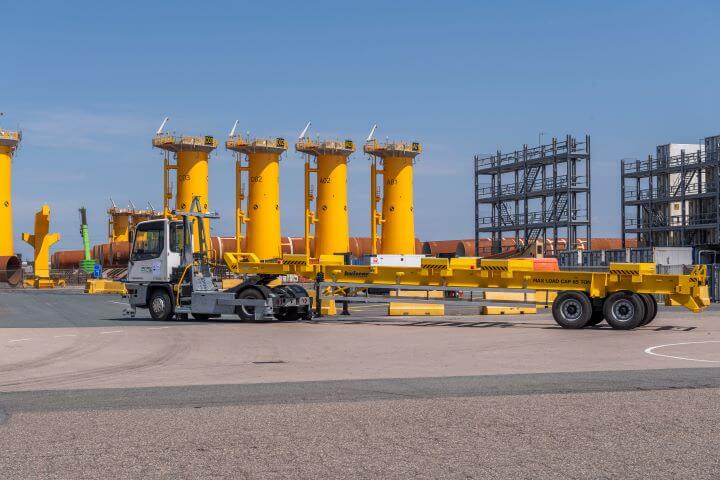 A Terberg autonomous terminal tractor is shown towing a yellow flatbed trailer at an industrial port area, with large yellow cylindrical structures in the background under a clear blue sky.
