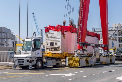 An autonomous terminal tractor by Terberg, equipped with sensor technology, is positioned under a towering red gantry crane handling a white shipping container, showcasing advanced logistics automation at a busy port.